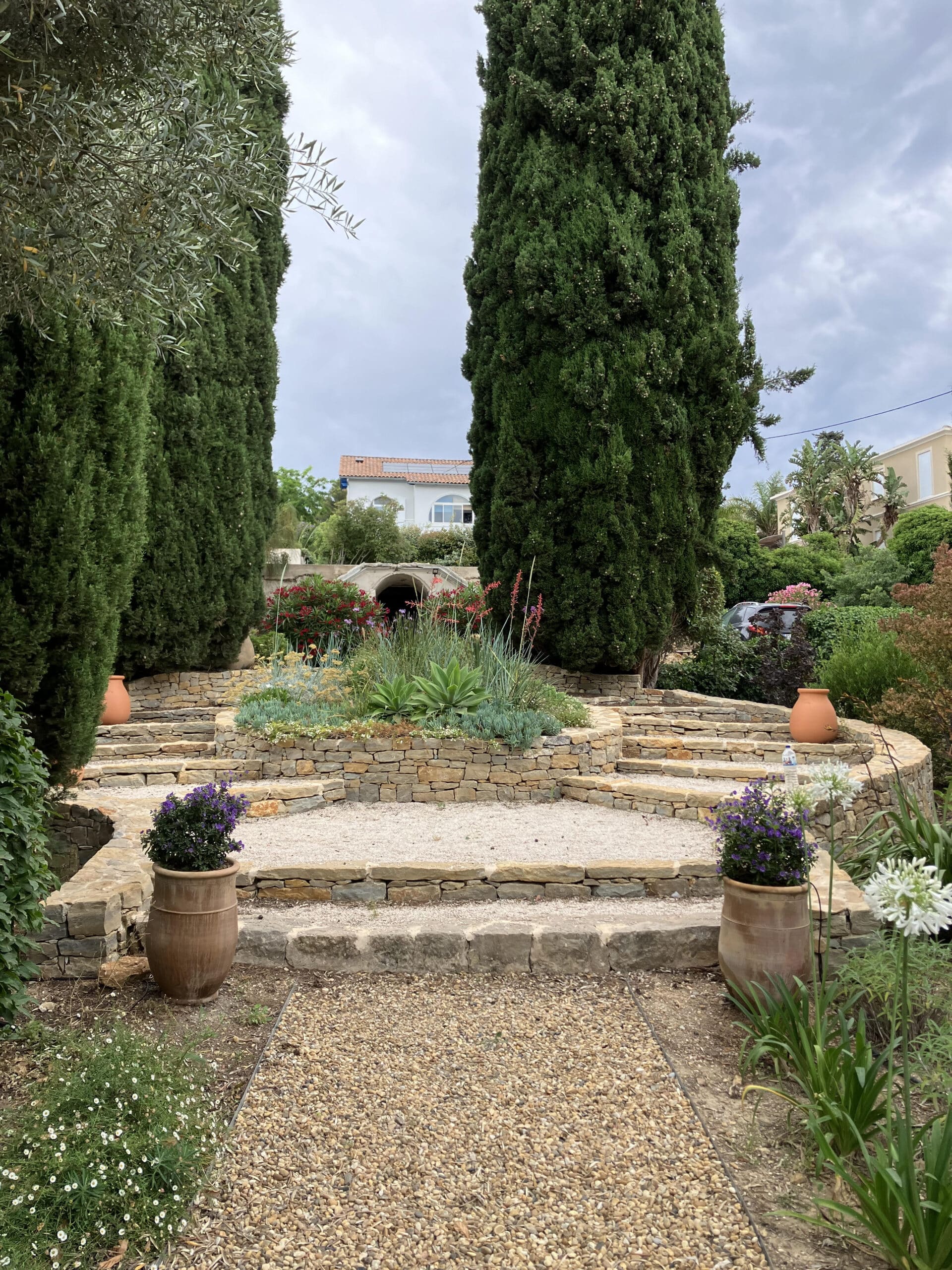 Jardin méditerranéen avec terrasses en pierre, cyprès élancés, pots en terre cuite et fleurs colorées sous un ciel voilé.