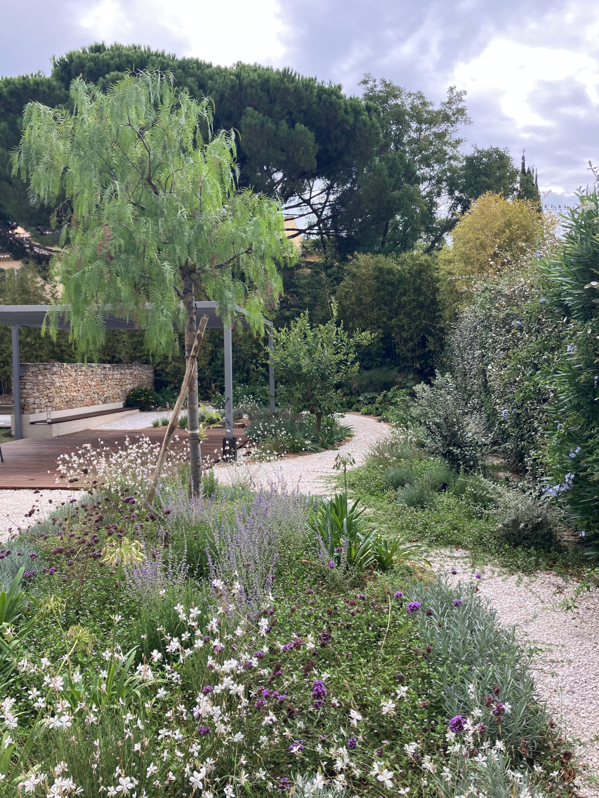 Jardin paysager avec chemin en gravier, fleurs sauvages, terrasse en bois, mur en pierre et pergola moderne.