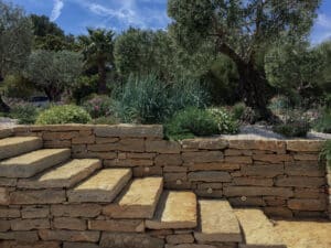 Escalier en pierre naturelle et mur de soutènement dans un jardin méditerranéen ensoleillé avec oliviers et palmiers.