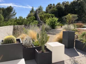 Jardin moderne avec cactus dans des pots gris, muret de pierre et végétation méditerranéenne sous un ciel bleu.