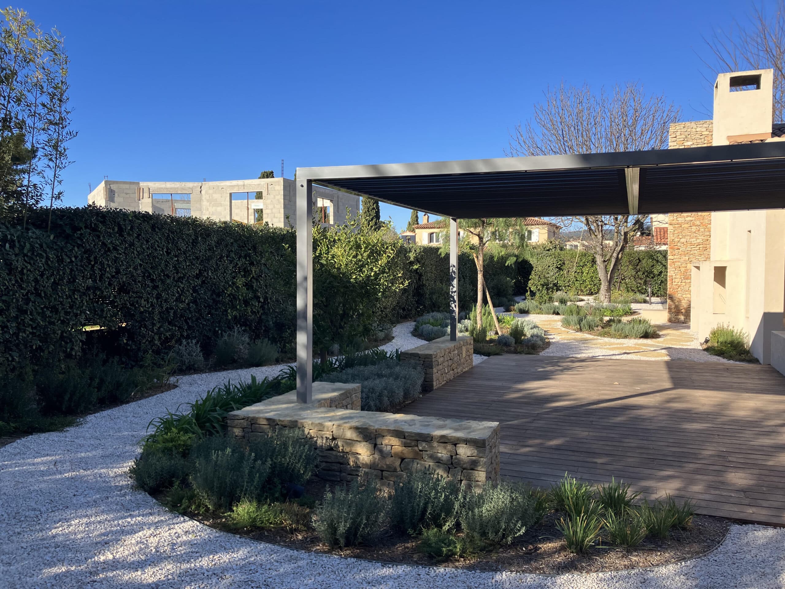 Pergola moderne sur terrasse en bois, muret en pierre et jardin paysager avec allées de gravier blanc sous ciel bleu.
