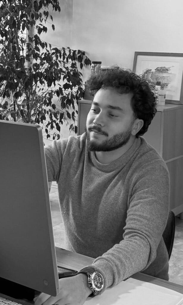 Travail sur ordinateur au bureau par un homme barbu Homme barbu travaillant sur ordinateur, portant un pull et une montre, fond de bureau avec plante.